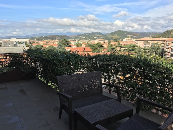 Eine Terrasse mit Korbmöbeln und Blick auf eine Stadt mit roten Dächern und grünen Hügeln im Hintergrund.