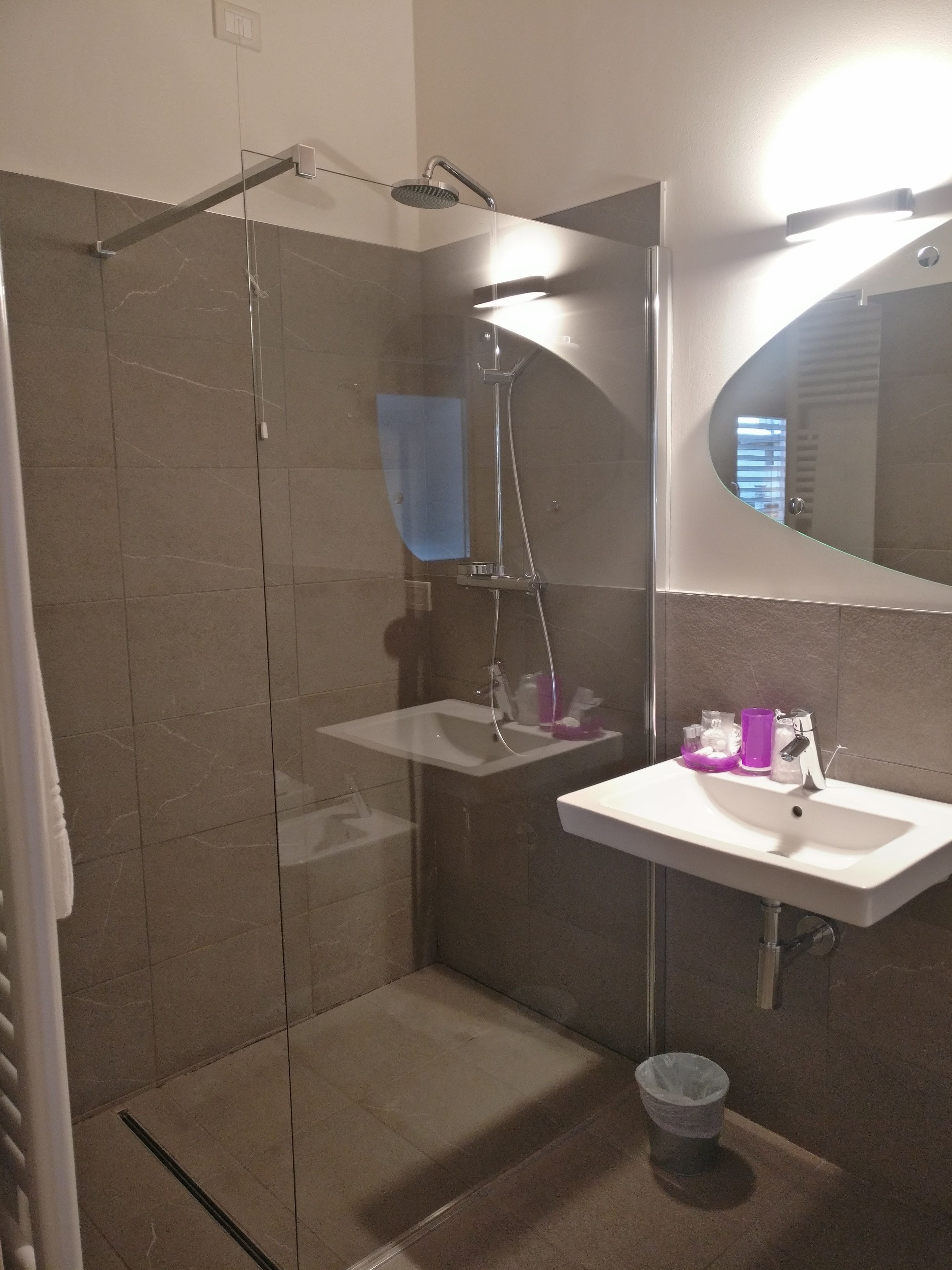 Une salle de bain moderne avec une douche à parois de verre, un lavabo blanc avec un miroir au-dessus et une petite poubelle.