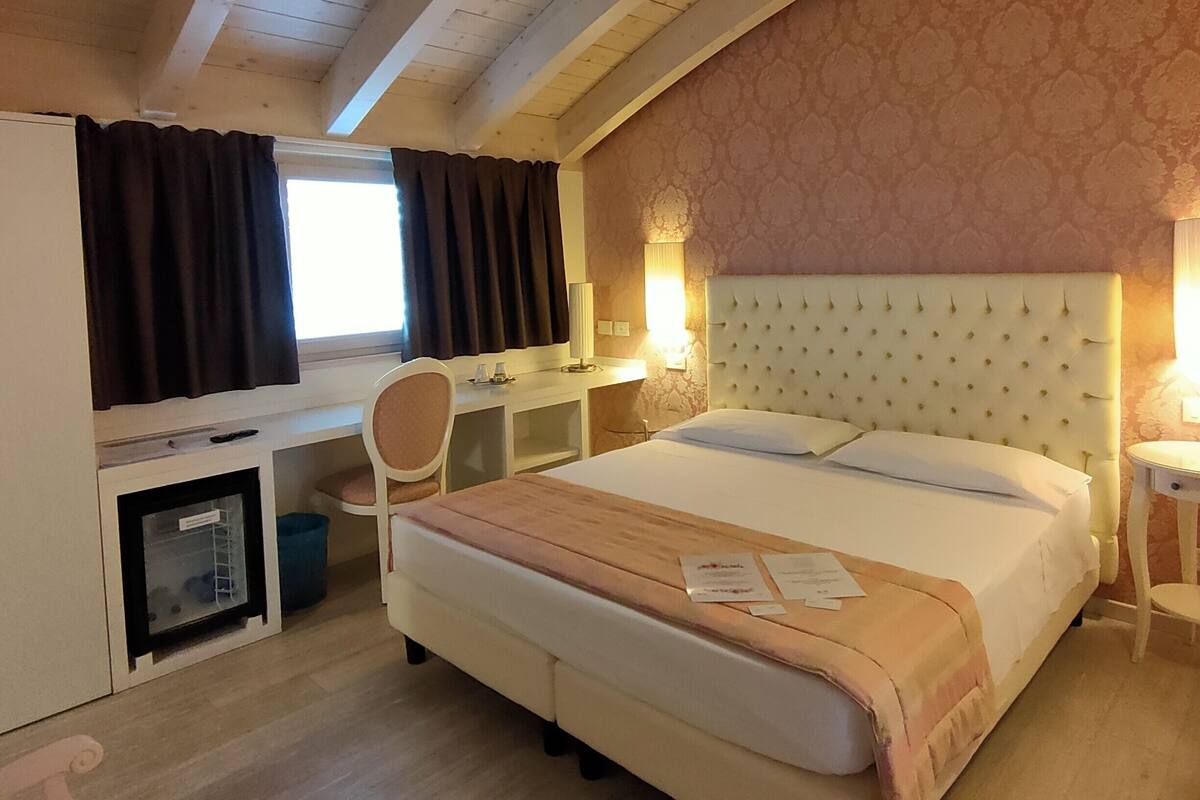 Habitación de hotel con una cama grande, un escritorio con silla, un televisor pequeño y una ventana con cortinas.