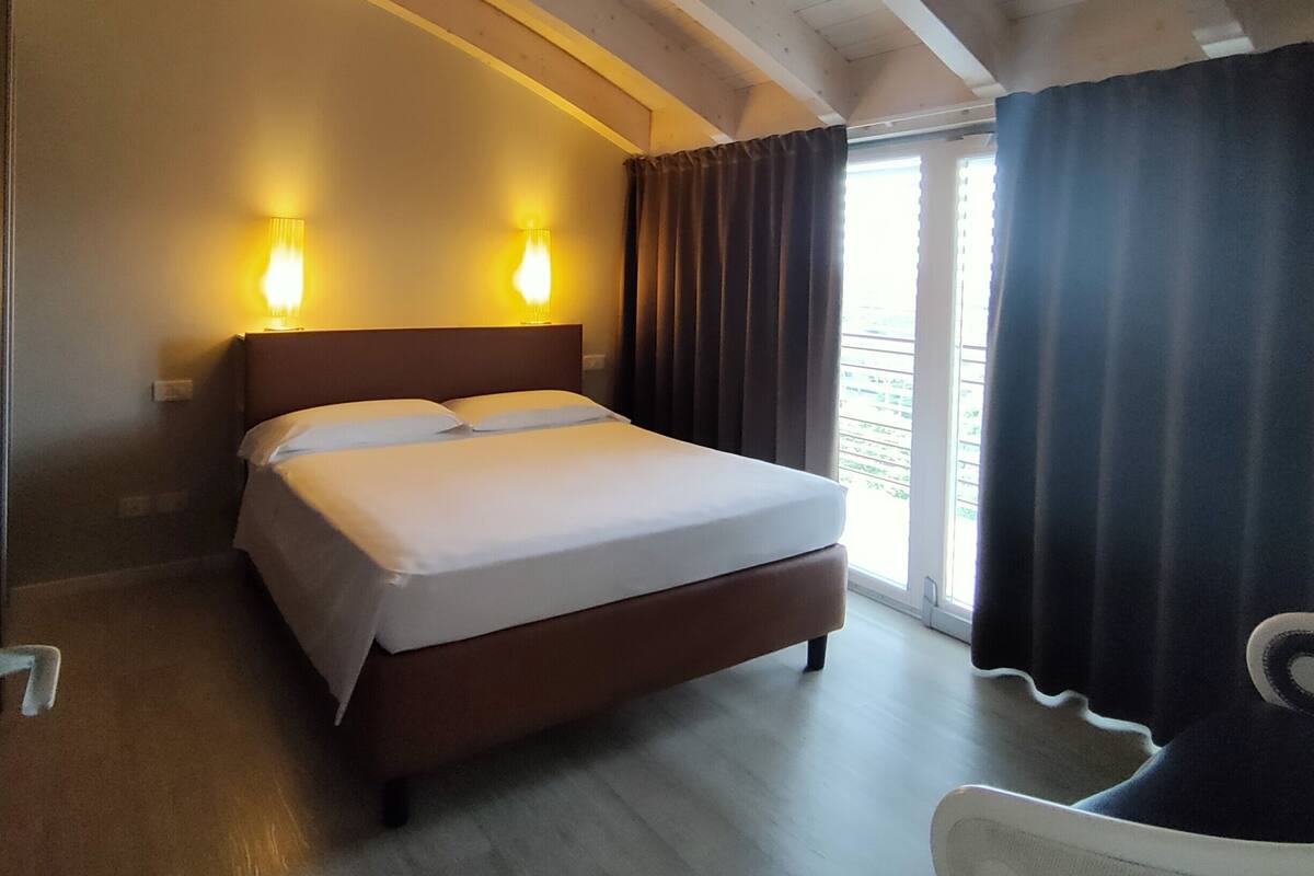 Habitación de hotel con cama, dos lámparas de noche, una silla y un ventanal con cortinas.