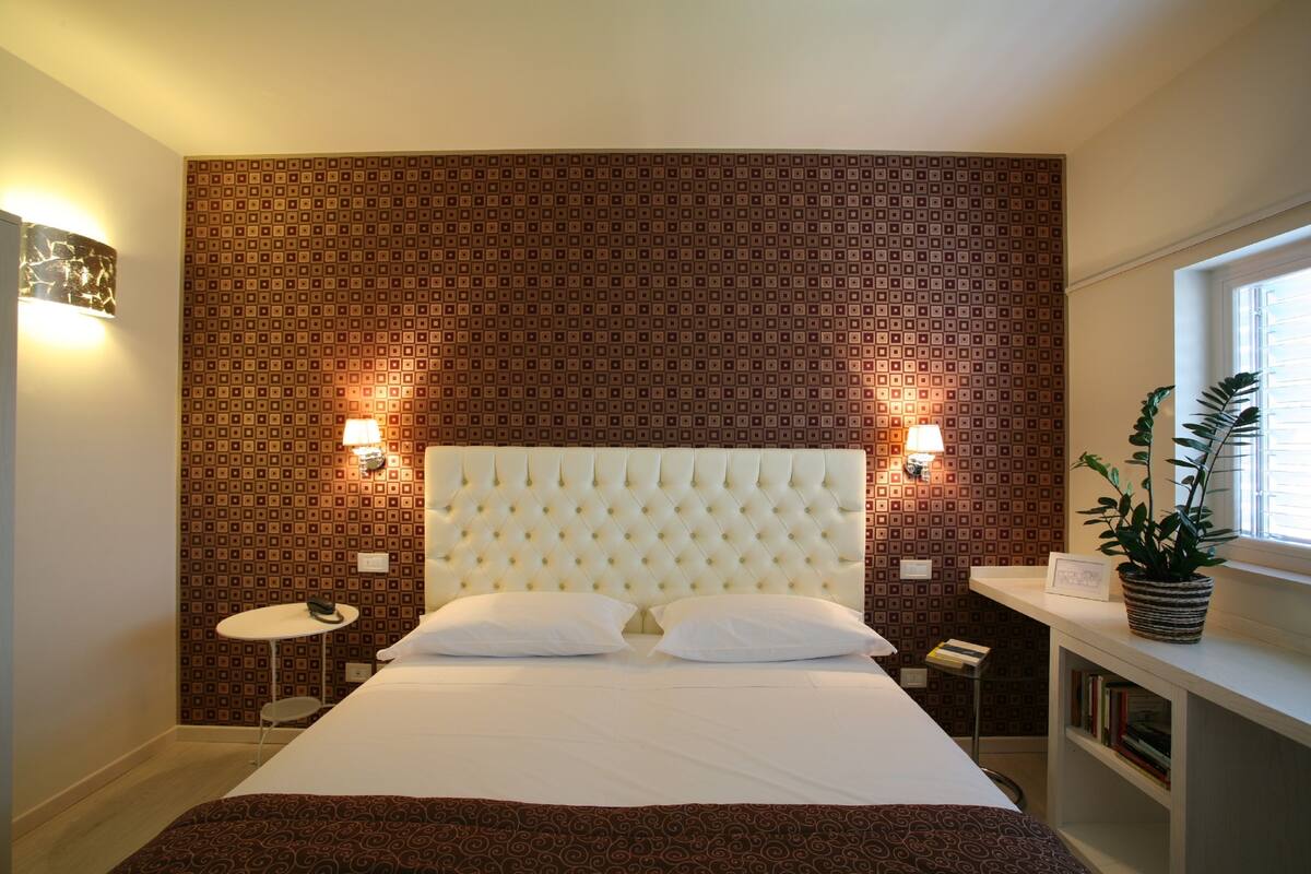 Habitación de hotel ordenada con una cama grande, dos lámparas de noche, una mesita redonda y una planta en maceta junto a la ventana.