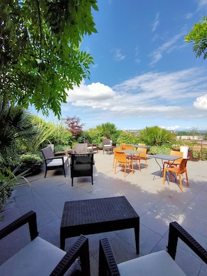 Ein Terrassenbereich im Freien mit Tischen und Stühlen, umgeben von Grünpflanzen und blauem Himmel.