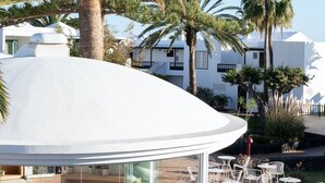 A modern outdoor seating area with round tables and chairs, surrounded by palm trees and a white building with a curved roof. - Barcarola Club (Tías)