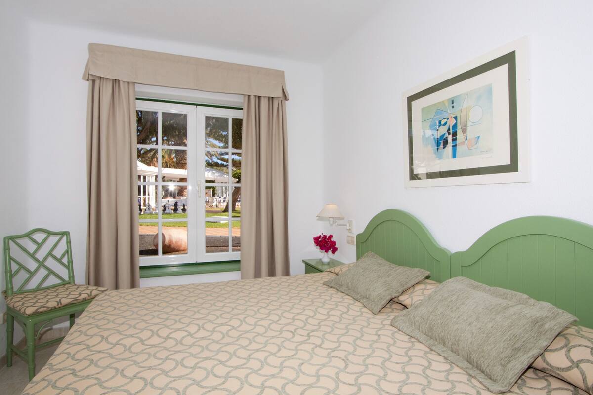 A hotel room with a bed, a chair, a window with curtains, a nightstand, a lamp, and a framed picture on the wall.