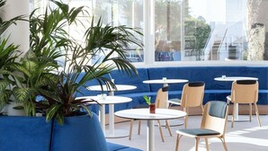 A modern lounge area with a blue sofa, round tables, and chairs, large windows with a view of palm trees and buildings. - Barcarola Club (Tías)