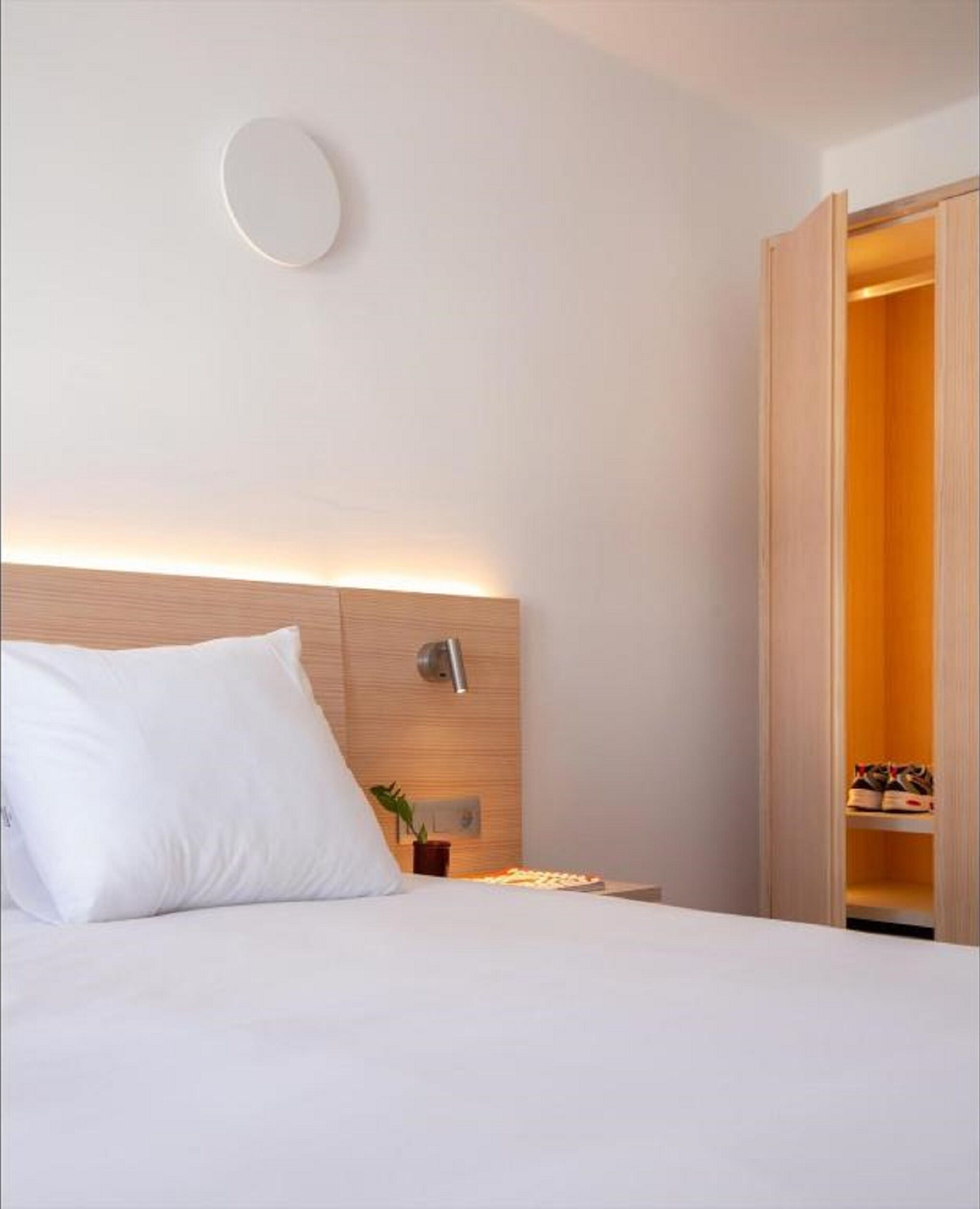 A hotel room with a bed, a bedside table with a lamp, a wall-mounted light, and a wooden wardrobe.