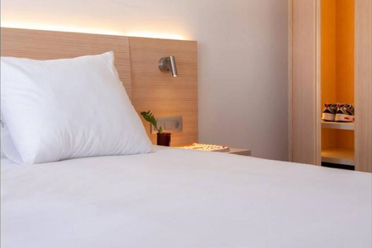 A hotel room with a bed, a bedside table with a lamp, a wall-mounted light, and a wooden wardrobe.