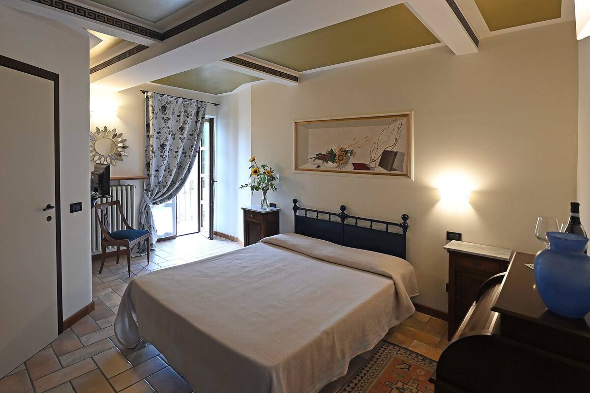 Une chambre à coucher avec un lit, une chaise, une table de chevet, un vase et un tableau accroché au mur.