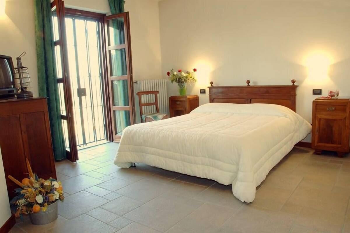 Une chambre avec un grand lit, une tête de lit en bois, une table de chevet, une commode, un radiateur, une fenêtre avec des rideaux et une vue sur l’extérieur.