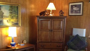 Un salon chaleureux avec des boiseries, un meuble en bois, une table recouverte d’une nappe en dentelle et un fauteuil agrémenté d’un coussin.