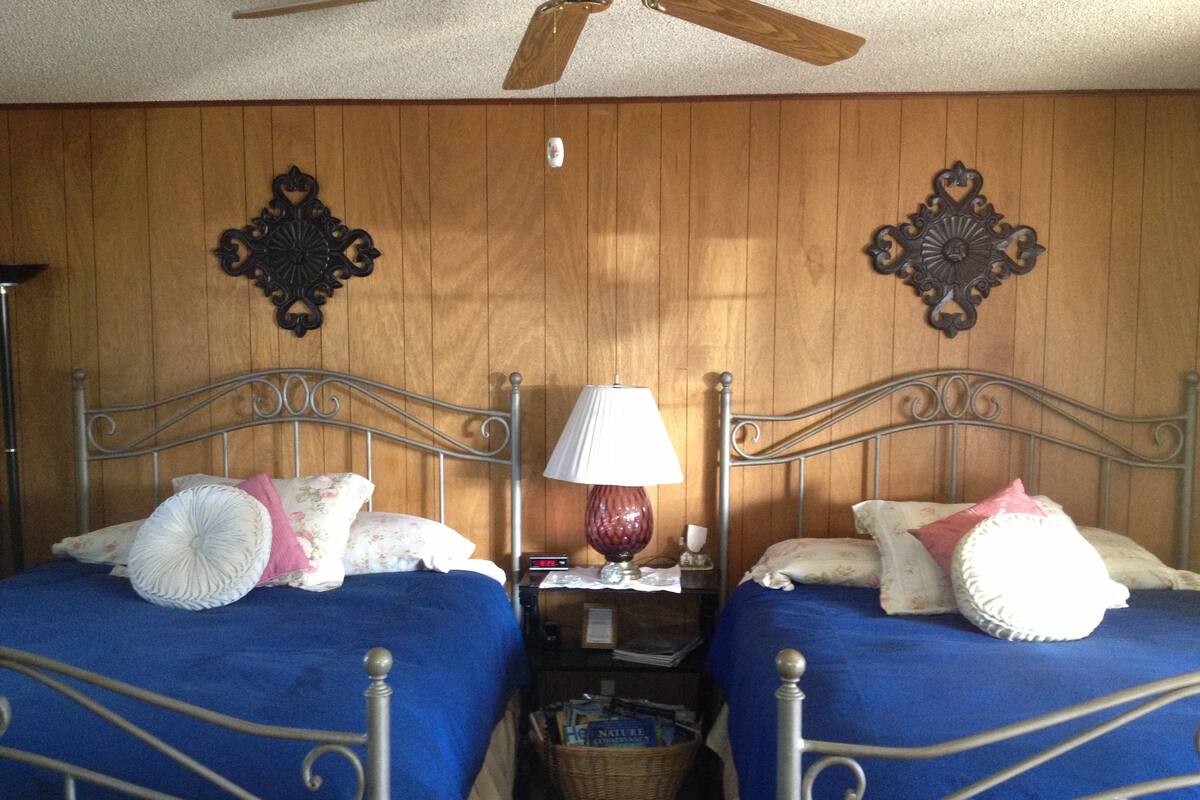Ein Schlafzimmer mit zwei Betten aus Metall, einem hölzernen Deckenventilator und einem Nachttisch mit Lampe und Korb.