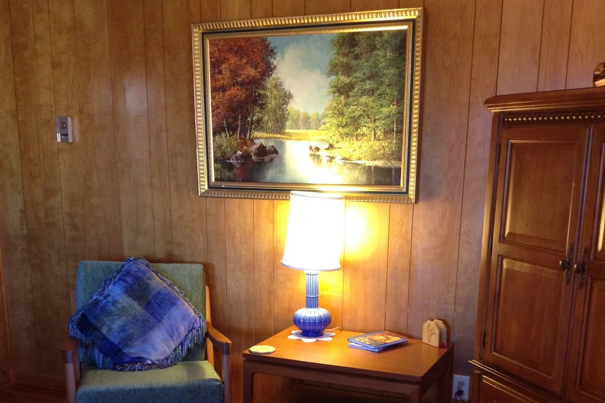 Ein Zimmer mit Holzwänden, ein Sessel mit blauer Sitzkissen, ein kleiner Tisch mit einer Lampe, ein gerahmtes Gemälde an der Wand und ein Holzschrank.