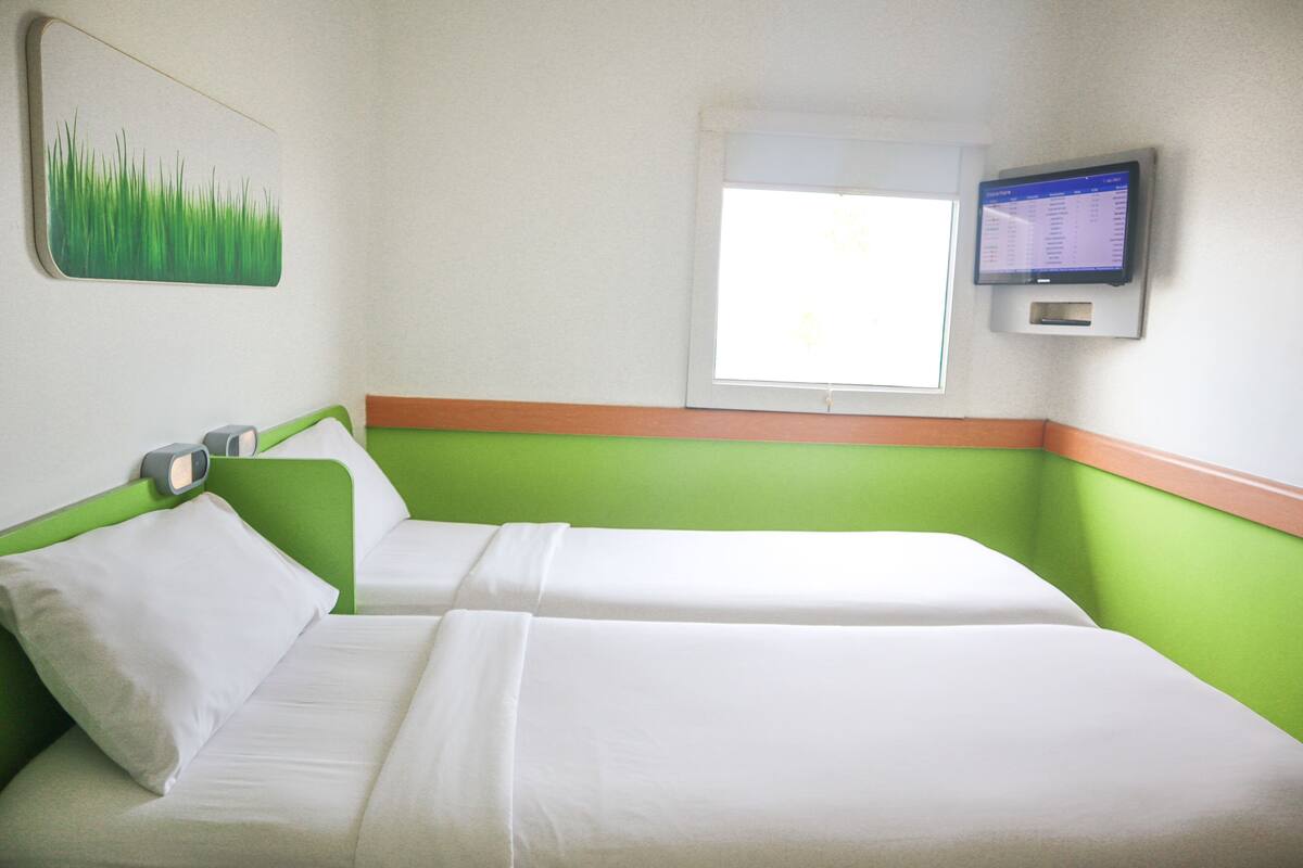 Ein Hotelzimmer mit zwei Betten, einem an der Wand montierten Fernseher und einem Fenster mit Jalousien.