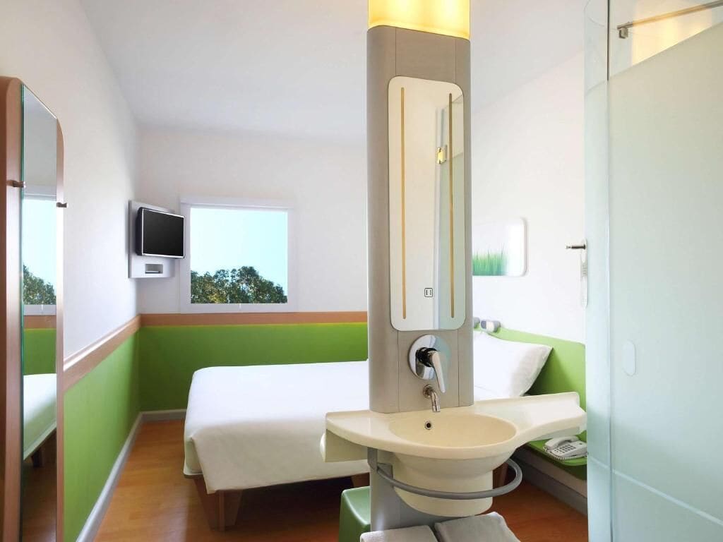 A hotel room with a bed, a sink, a mirror, a television, and a window with a view of trees.