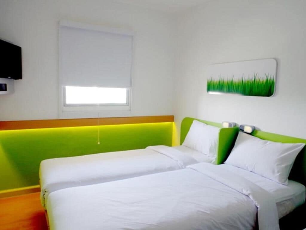 A hotel room with two single beds, a green headboard, a white wall, a window with blinds, and a flat-screen TV mounted on the wall.