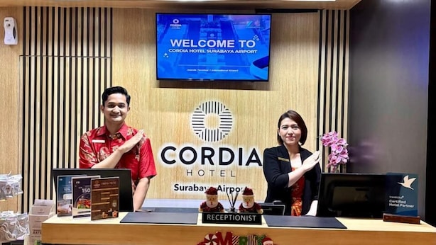 A reception desk at Cordia Hotel Surabaya Airport with a 'Welcome to Cordia Hotel Surabaya Airport' sign, a receptionist, and a staff member.