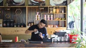 A barista preparing a drink at a coffee shop with shelves stocked with coffee beans and condiments.