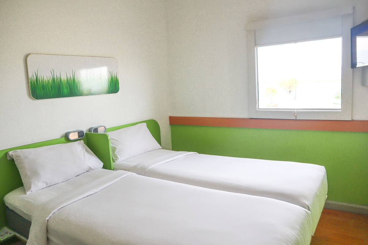 Ein Zimmer mit zwei Einzelbetten, einem grünen Kopfteil, weißer Bettwäsche, einem an der Wand hängenden Bild von Gras und einem Fenster mit Blick auf Grünflächen.