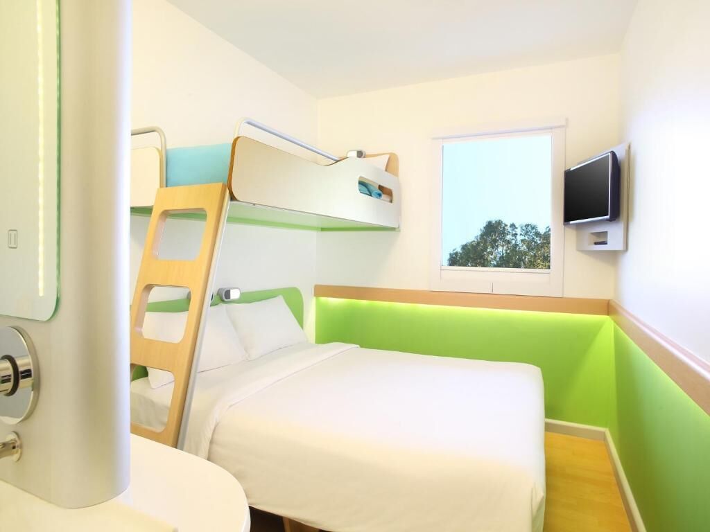 A compact hotel room with a bunk bed, a single bed, a television mounted on the wall, and a window with a view of trees.
