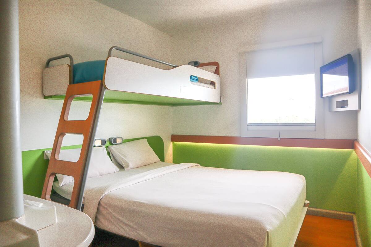 Ein Etagenbett mit einer Stehle, ein Einzelbett, ein kleiner Tisch und ein Fenster mit Jalousien.