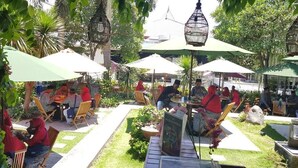 An outdoor seating area with tables and chairs under umbrellas, surrounded by greenery and a stone pathway.