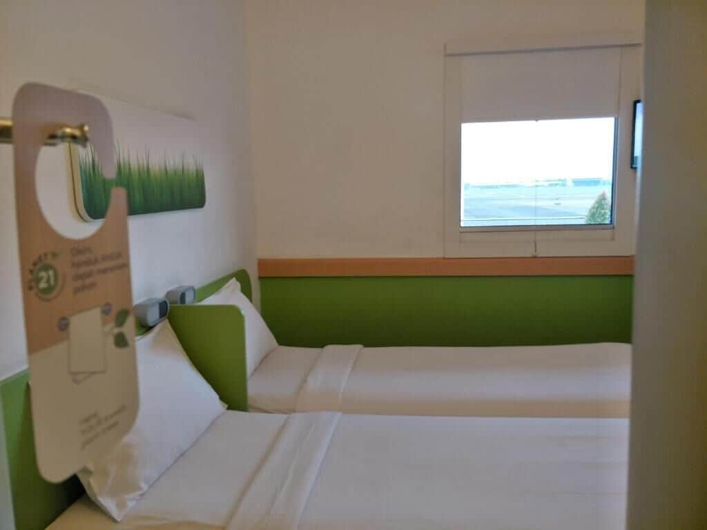 Een hotelkamer met twee bedden, een raam met uitzicht en een toiletrolhouder aan de muur.