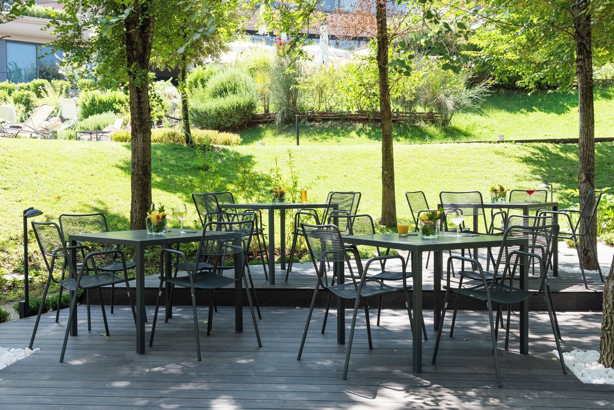 An outdoor patio with metal tables and chairs, surrounded by greenery and trees.