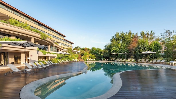 A modern hotel with a large, curved swimming pool surrounded by sun loungers and umbrellas.