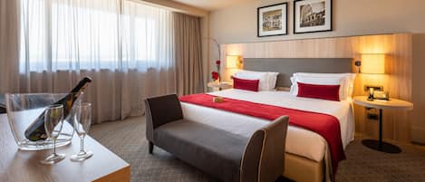 Quarto de hotel com uma cama, mesas de cabeceira, escrivaninha com cadeira, uma janela grande com cortinas e duas imagens emolduradas na parede.