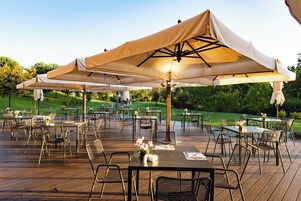 An outdoor dining area with tables and chairs under large umbrellas, surrounded by greenery.