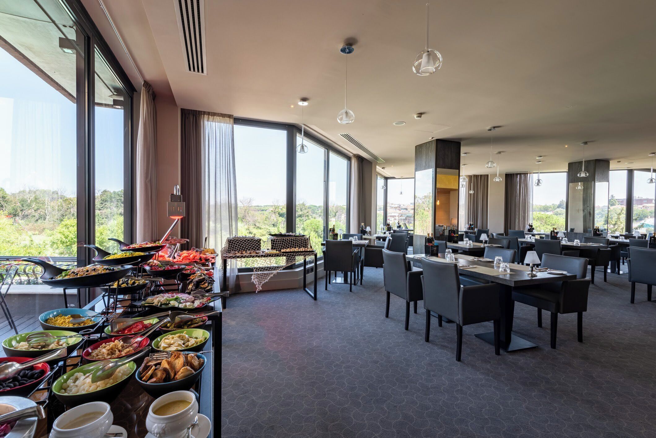 A spacious dining area with multiple tables and chairs, large windows offering a view of the outdoors, and a buffet with various food items.