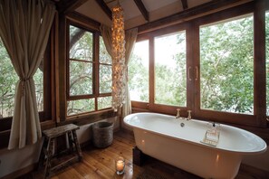 A bathroom with a freestanding bathtub, wooden flooring, a window with curtains, and a hanging light fixture.
