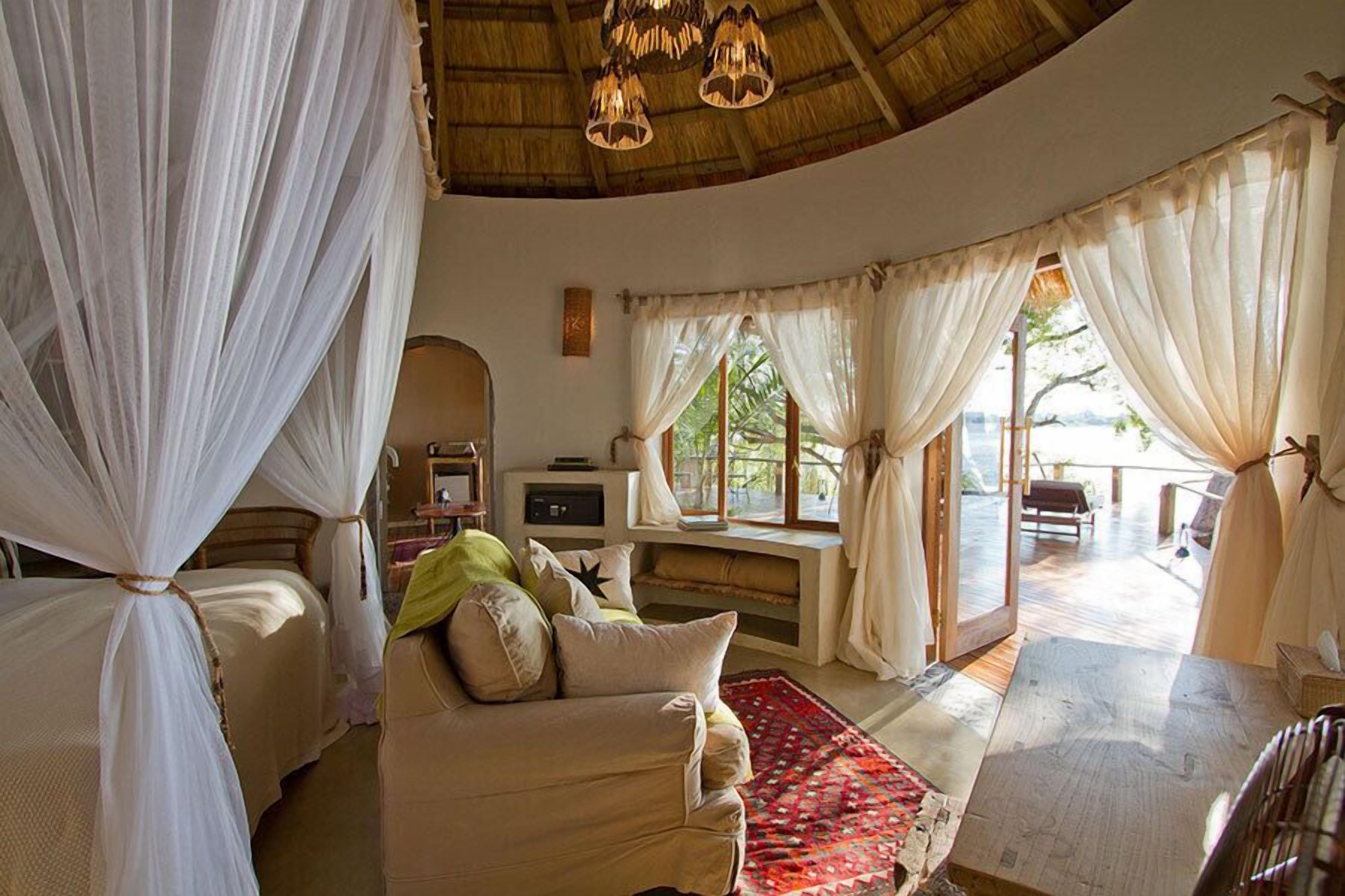 A cozy room with a bed, a sofa, and a table. There are curtains and a window with a view.