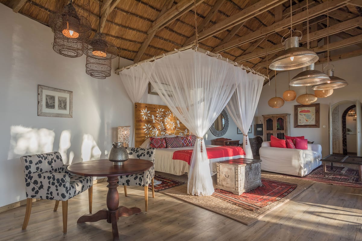 A spacious room with a wooden ceiling, a bed with a canopy, a dining table with chairs, and a large window allowing natural light.