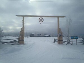 A wooden archway with a hanging ornament, leading to a snow-covered path and buildings in the background.