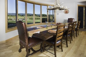 A dining room with a large table, wooden chairs, and a view of a green landscape through large windows.