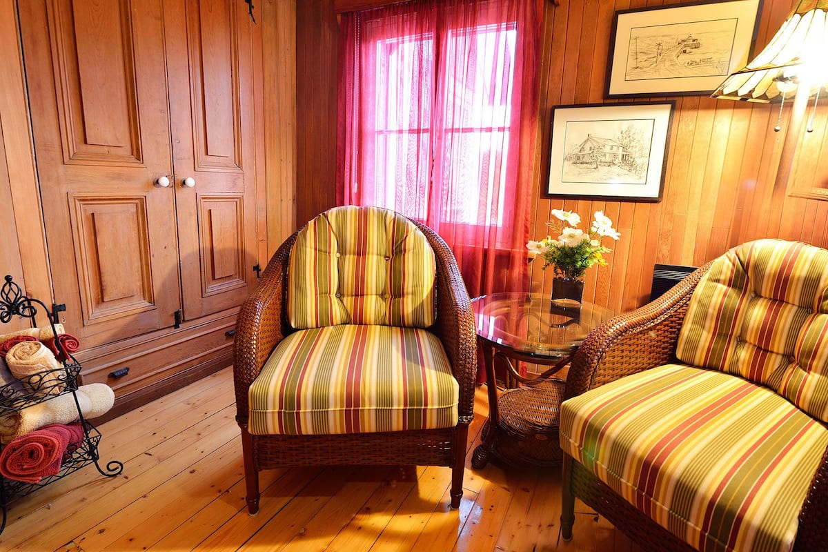 Una habitación acogedora con paneles de madera, un sillón a rayas, una silla de mimbre, una mesita con un florero y un dibujo enmarcado en la pared.