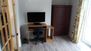 Una habitación con un escritorio de madera, una silla de oficina negra, un televisor en la pared, un armario y una ventana con cortinas.