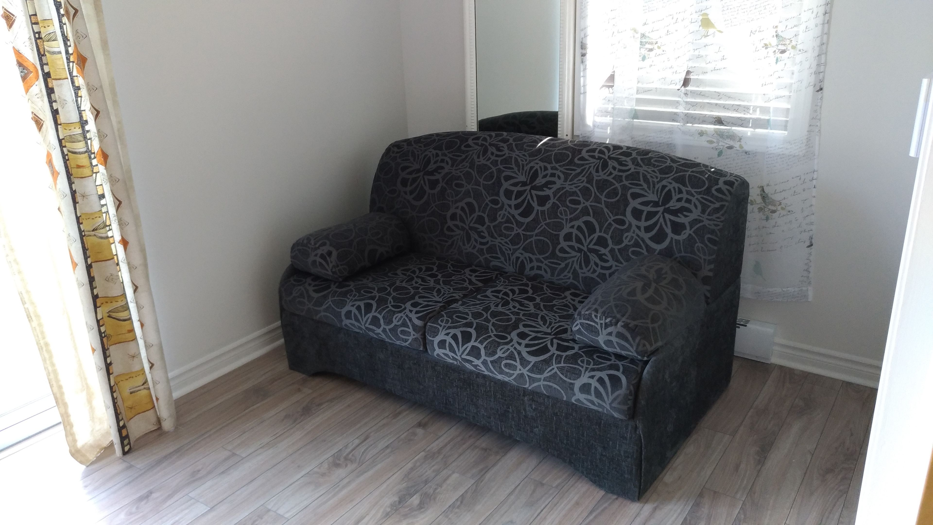 A grey patterned sofa in a room with wooden flooring and a window with a curtain.
