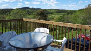 A deck with a glass-top table and white chairs, overlooking a green landscape with hills and a red car in the background. - Les Appartements de la Bergeronnette (Les Bergeronnes)