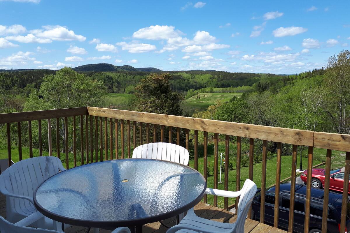 A deck with a glass-top table and white chairs, overlooking a green landscape with hills and a red car in the background.