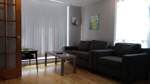 A living room with a dark grey sofa, a wooden coffee table, and a small black table with a green object on it. There is a window with blinds, a painting on the wall, and a door with a wooden frame. - Les Appartements de la Bergeronnette (Les Bergeronnes)