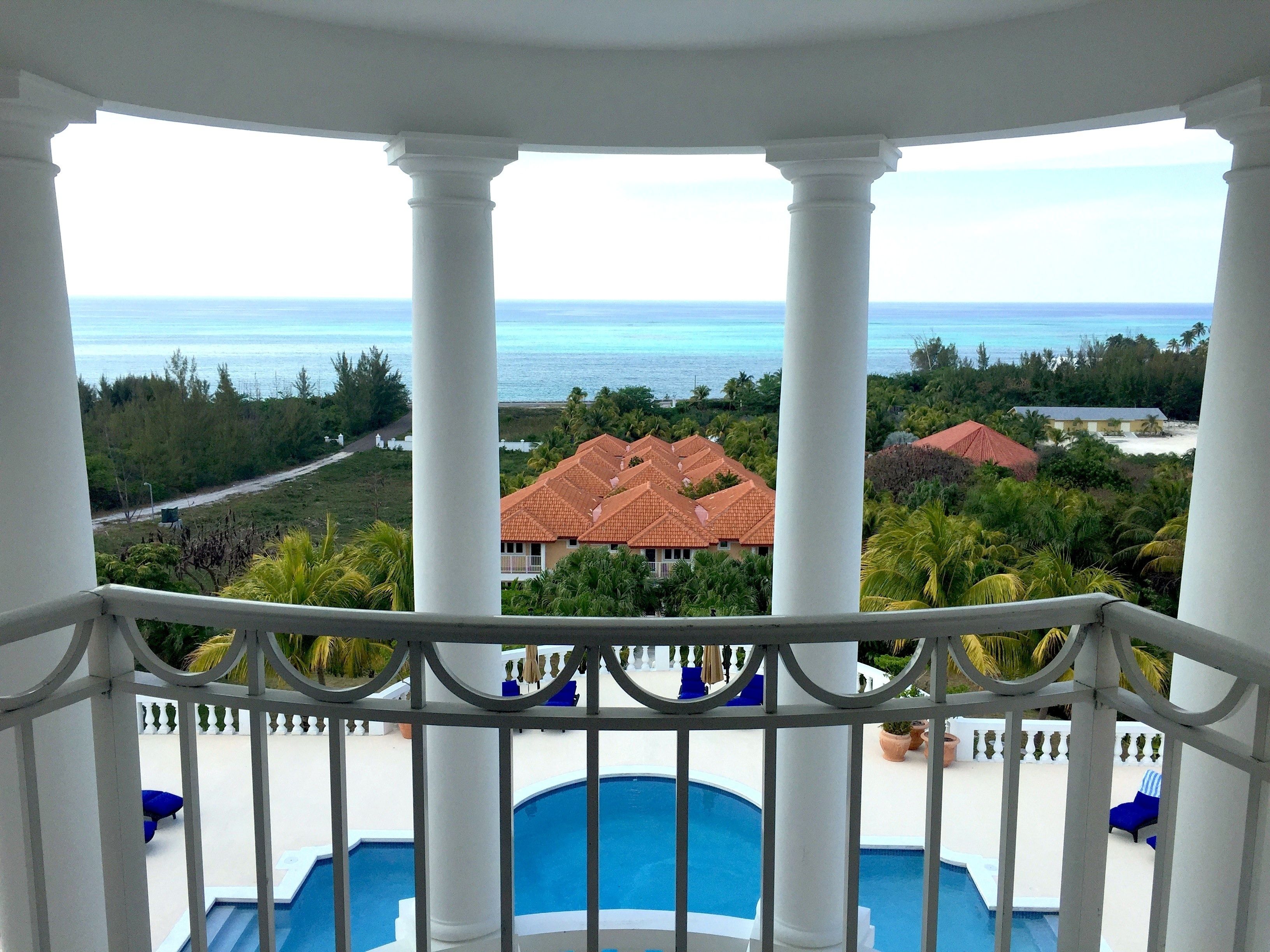 A balcony with a pool, white columns, and a view of the ocean and resort buildings.