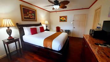 A hotel room with a bed, a ceiling fan, a painting, a lamp, a chair, a desk with a TV, and a door.