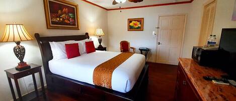 A hotel room with a bed, a ceiling fan, a painting, a lamp, a chair, a desk with a TV, and a door.