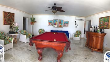 A room with a pool table, white wicker chairs, a wooden cabinet, and wall art.