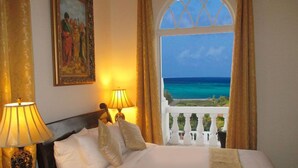Habitación de hotel con cama, dos lámparas de noche, un cuadro en la pared y vista al océano a través de una ventana con balcón.