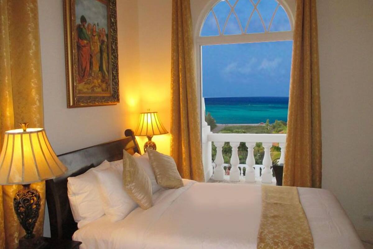 Een hotelkamer met een bed, twee bedlampen, een schilderij aan de muur en uitzicht op de oceaan door een raam met balkon.