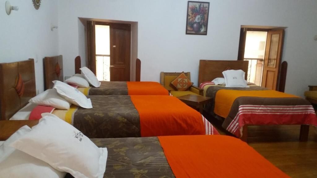 Habitación de hotel con tres camas individuales, cada una con una colcha de color diferente. Se observan cabeceros de madera y una pequeña mesa con una silla en el centro.