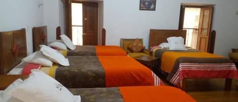 Habitación de hotel con tres camas individuales, cada una con una colcha de color diferente. Se observan cabeceros de madera y una pequeña mesa con una silla en el centro.